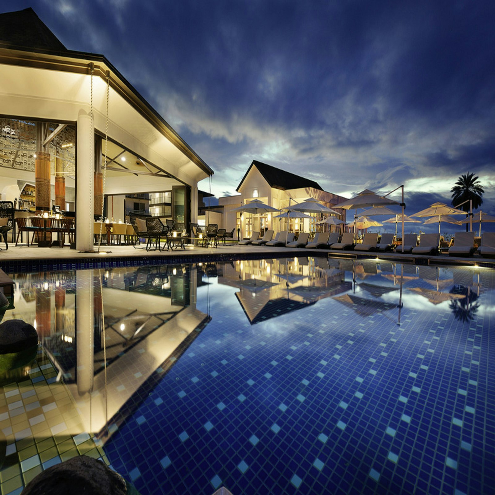 Palm-lined resort swimming pool glowing at dusk beside a candlelit terrace